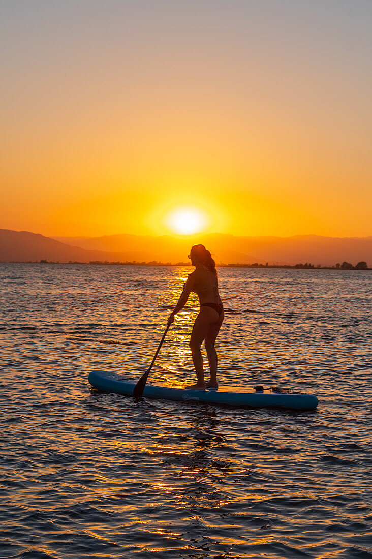 Silhouette einer jungen Frau beim Paddelsurfen während des Sonnenuntergangs am Strand von Trabucador, Ebro-Delta, Tarragona, Spanien