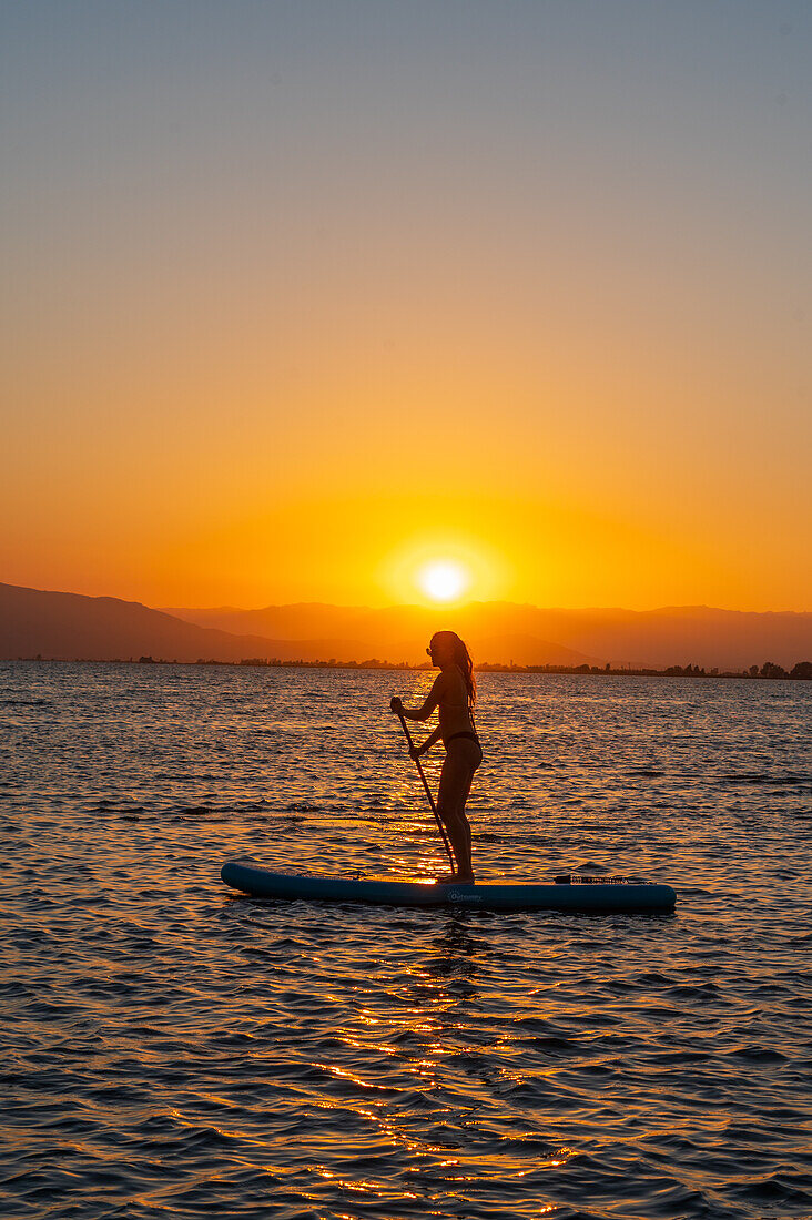 Silhouette einer jungen Frau, die bei Sonnenuntergang am Strand von Trabucador, Ebro-Delta, Tarragona, Spanien, Paddelsurfen übt