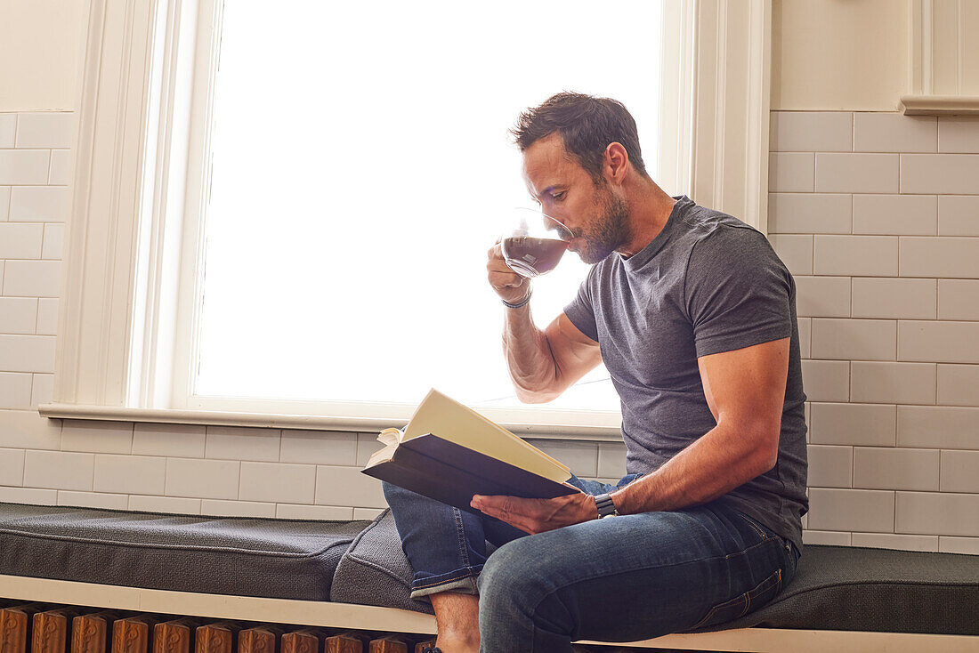 Man reading book and drinking tea by window at home\n