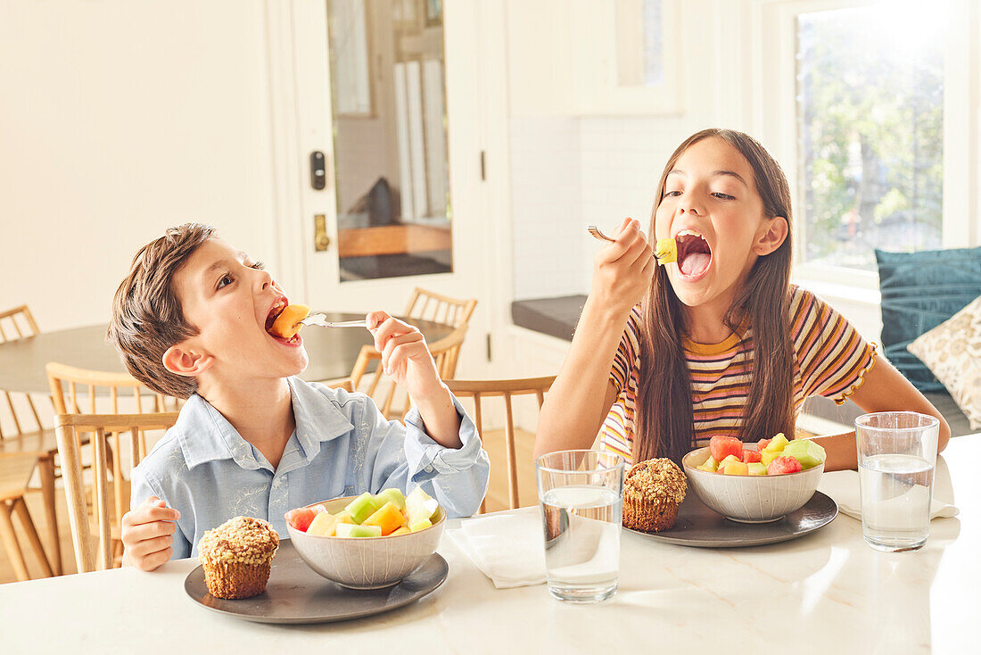 Boy (8-9) and girl (12-13) eating breakfast in kitchen\n