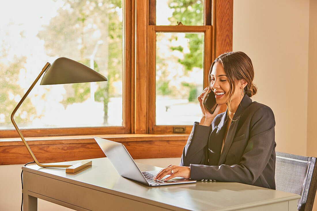 Lächelnde Frau mit Laptop und Smartphone am Schreibtisch zu Hause