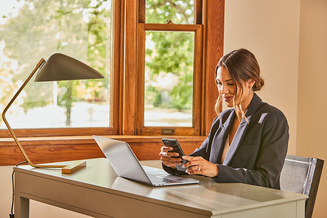 Lächelnde Frau mit Laptop und Smartphone am Schreibtisch zu Hause