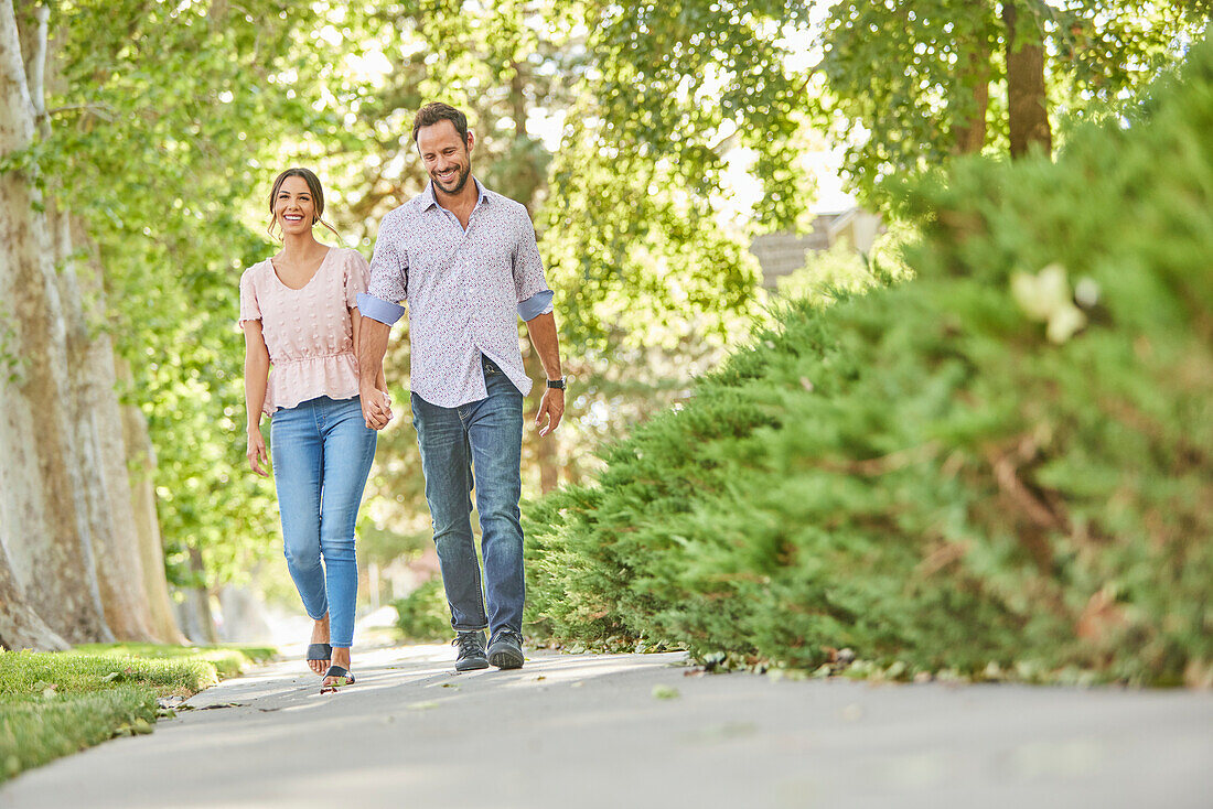 Smiling couple holding hands, walking on treelined sidewalk\n