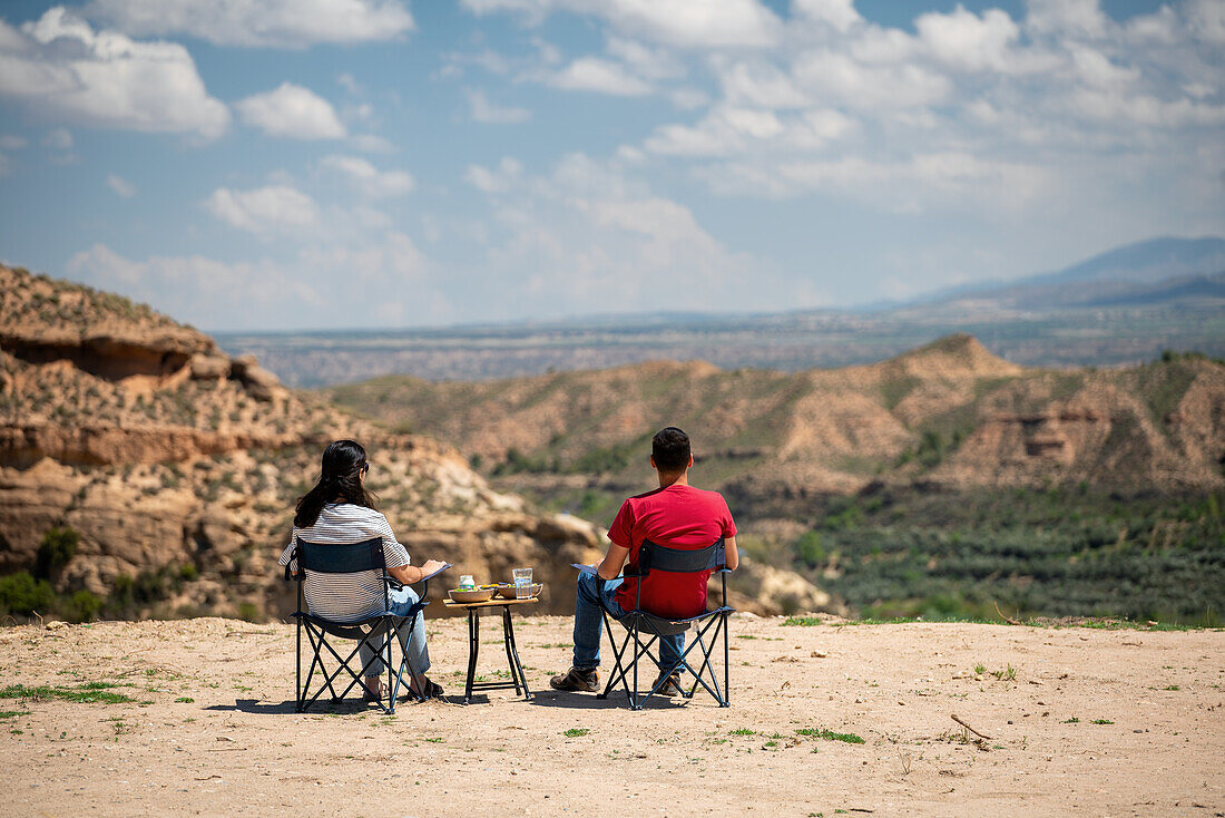 Paar sitzt auf Stühlen mit Tisch und blickt auf eine wüstenartige Landschaft in der Nähe des Francisco Abellan-Staudamms, Granada, Andalusien, Spanien, Europa