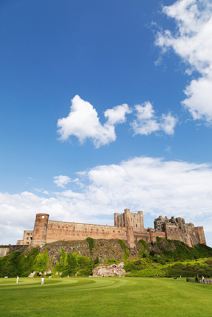 Bamburgh Castle, eine mittelalterliche Festung, denkmalgeschütztes Gebäude auf einem zerklüfteten Felsvorsprung aus vulkanischem Dolerit, mit Blick auf einen Cricketplatz, Bamburgh, Northumberland, England, Vereinigtes Königreich, Europa