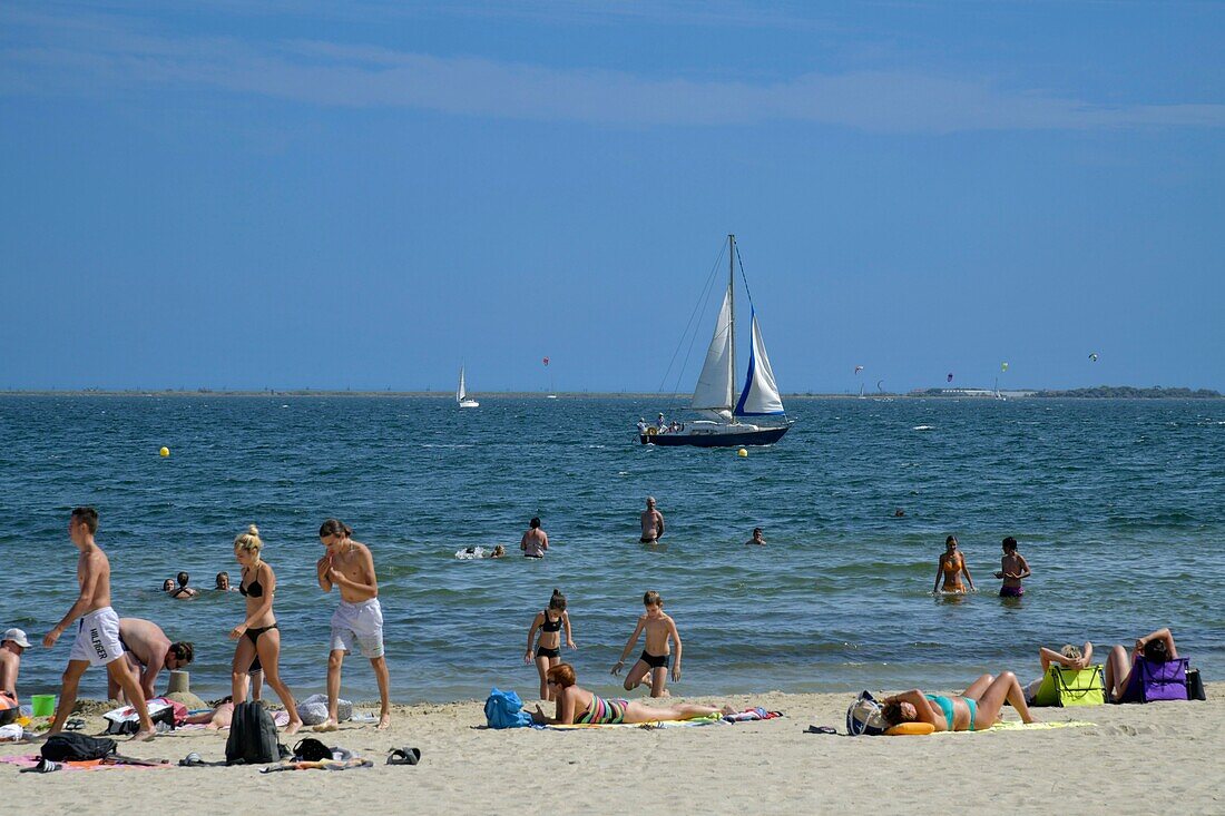 Frankreich, Herault, Meze, Urlauber an einem Strand in der Lagune von Thau mit einem Segelboot im Hintergrund