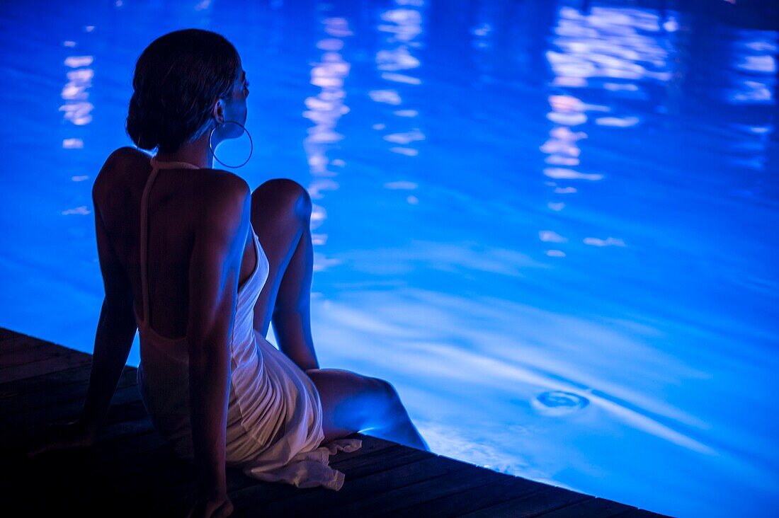 France, Caribbean, Lesser Antilles, Guadeloupe, Grande-Terre, Le Gosier, a woman relaxes at night by the swimming pool of the Creole Beach Hotel\n