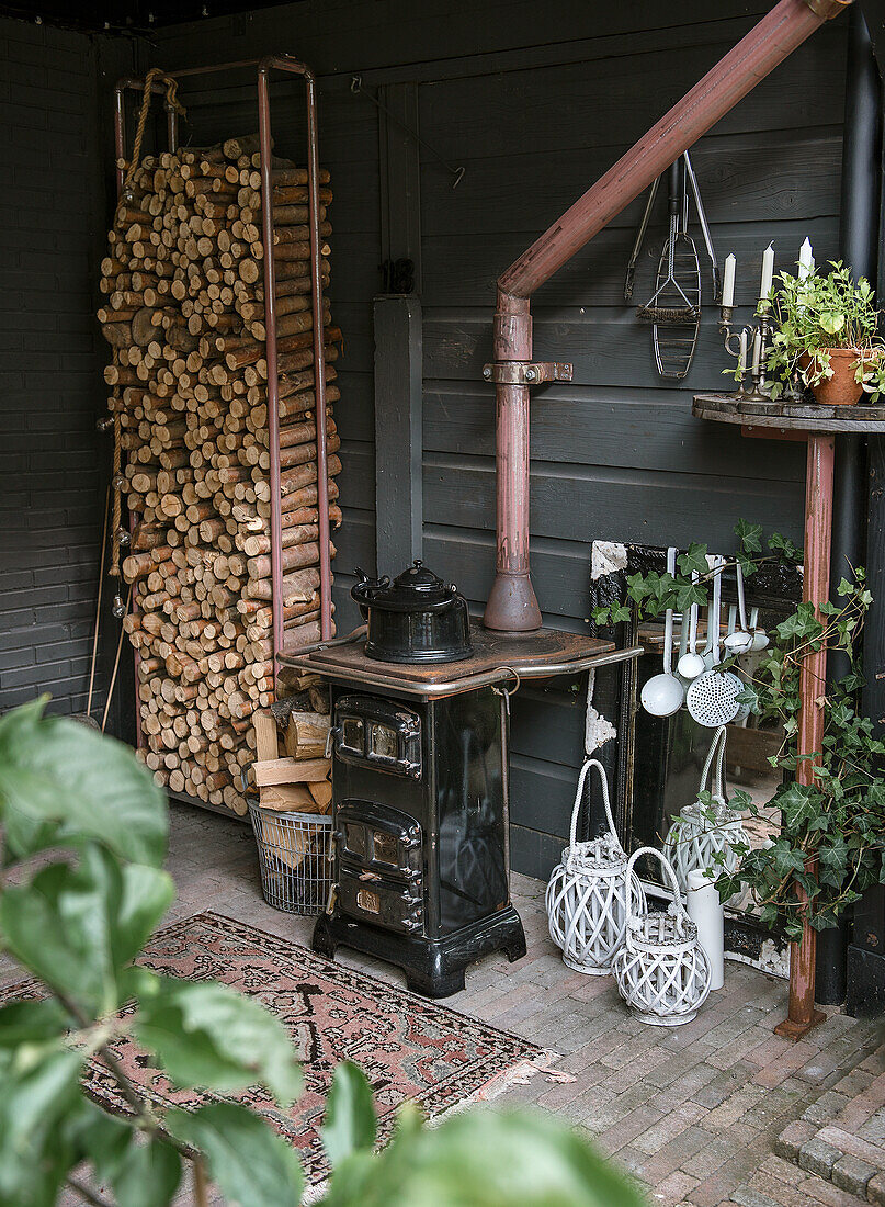 Holzstapel und nostalgischer Ofen in rustikaler Ecke mit Laternen und Pflanzen