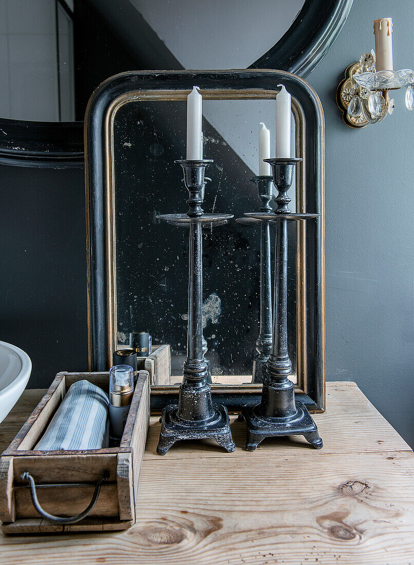 Black candlesticks with white candles in front of an antique mirror on a wooden chest of drawers