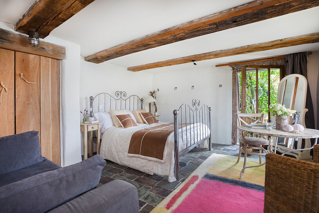 Country-style bedroom with wooden beamed ceiling and iron bed, seating area by the window