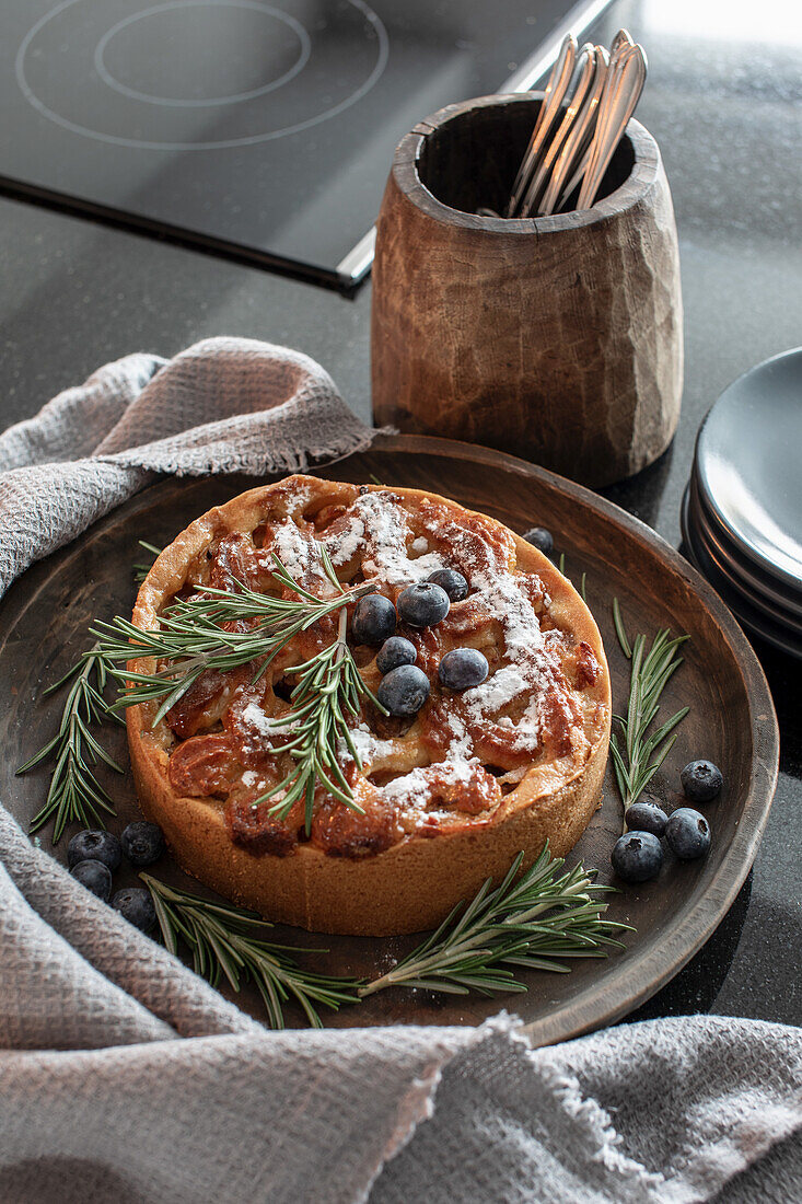 Apfelkuchen mit Rosmarinzweigen und Heidelbeeren auf Holzplatte