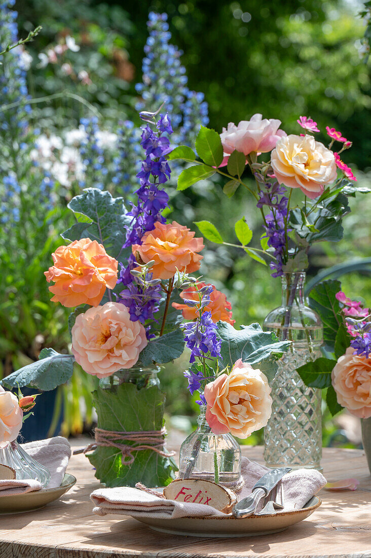 Kleinstrauchrose 'Cubana' (Rosa), Beetrose 'Tequila' und Rittersporn, einzelne Blüten in Vasen auf gedecktem Gartentisch