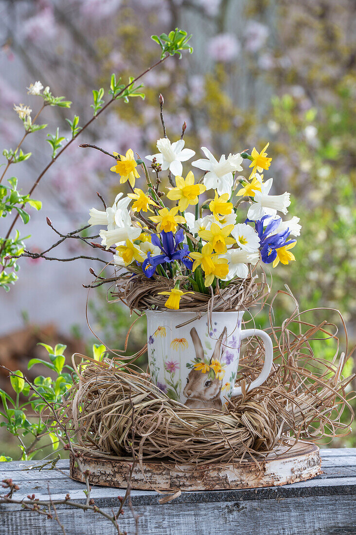 Narzissen (Narcissus) 'Sailboat' ,'Tete a Tete', Zwergiris (Iris Reticulata )'Harmony' in Vase mit Osternest aus Stroh