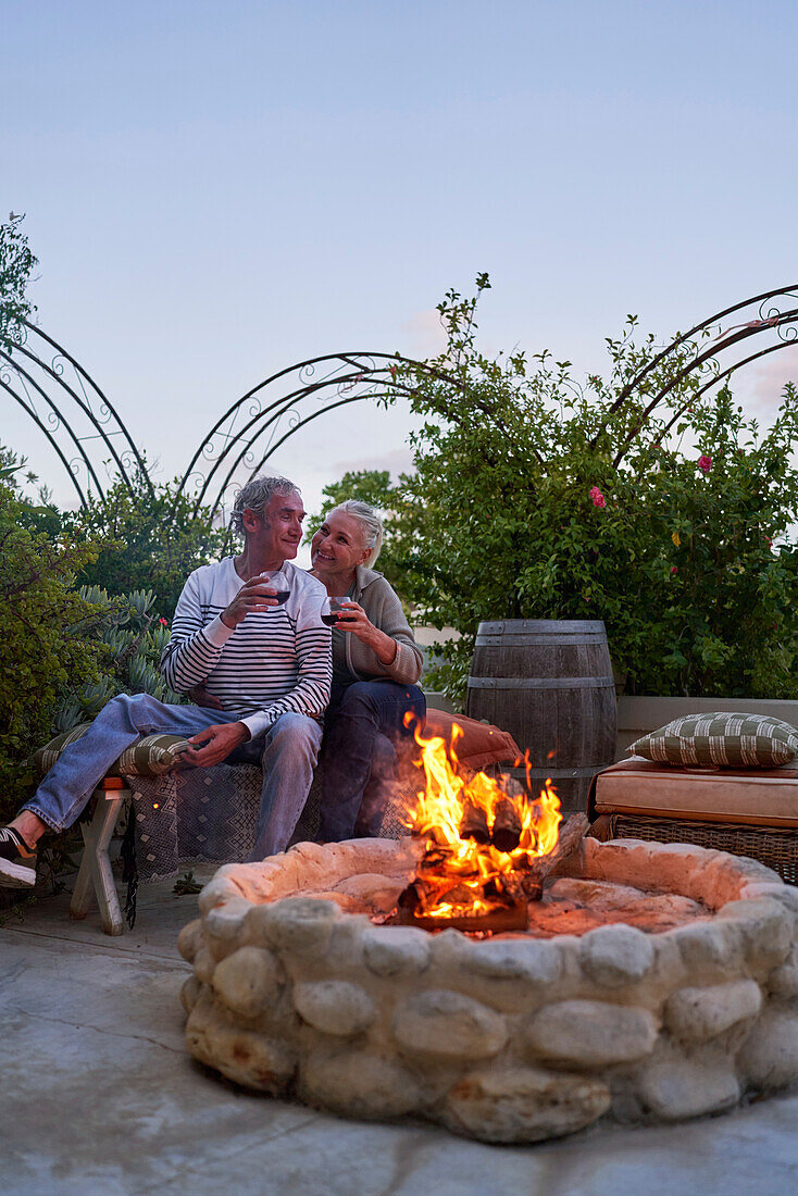 Senior couple drinking wine by fire pit on summer patio