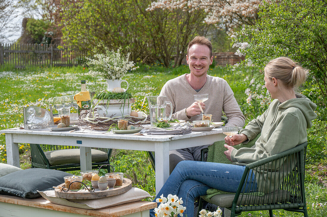 Young couple sitting at a set table for Easter breakfast with Easter nest, colored eggs, cereal bowls and water jug, and bouquet of flowers in the garden