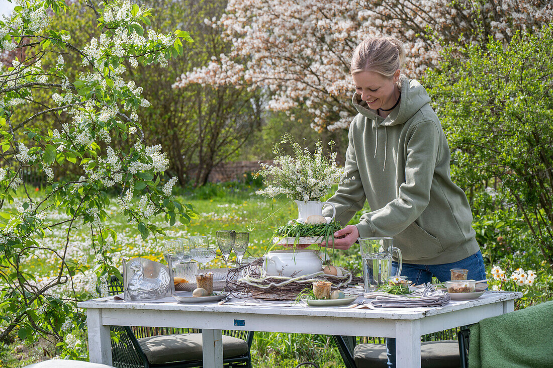 Junge Frau stellt Blumenstrauß auf gedeckten Tisch zum Osterfrühstück mit Osternest und gefärbten Eiern, vor blühenden Sträuchern
