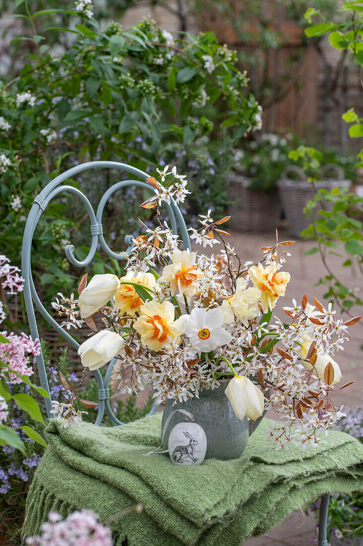 Blumenstrauß aus Felsenbirne, Tulpen, Narzissen 'Poeticus' und 'Tahiti' in Vase mit bemaltem Osterei auf Gartenstuhl