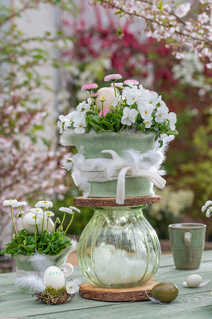 Etagere mit Blumentopf aus Hornveilchen, Gänseblümchen und Glasvase, Ostereier in kleinem Moosnestern mit Federn und auf Tisch