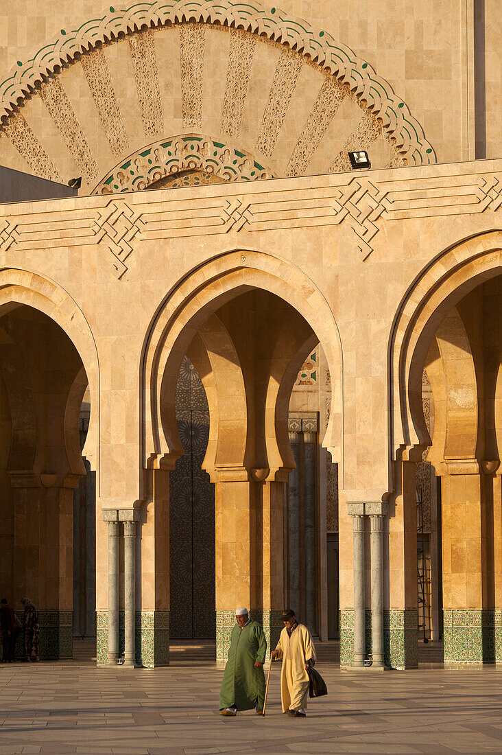 Morocco,Two men walking across courtyard in front of Hassan II mosque at dusk,Casablanca