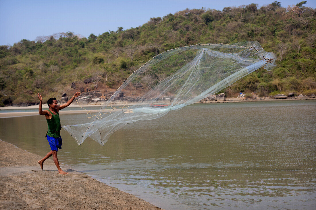 Ein einsamer Fischer mit einem einzigen Netz, das er am Strand von Palolem in das seichte Mündungswasser wirft, um kleine Fische zu fangen,Goa,Indien.