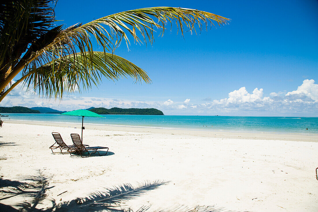 Malaysia,Pulau Langkawi,Liegestühle unter Sonnenschirm am weißen Sandstrand mit Palmen mit Blick auf das blaue Meer, Pantai Cenang (Cenang Strand)