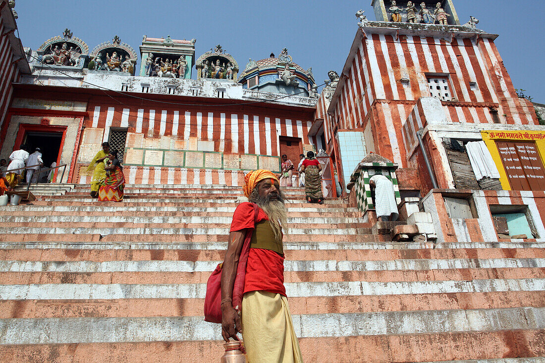 Hindu-Sadhu, der an einem Hindu-Tempel und den farbenfrohen Stufen des Bade-Ghat oberhalb des Ganges vorbeigeht. Die Kultur von Varanasi ist eng mit dem Fluss Ganges und seiner religiösen Bedeutung verbunden: Sie ist die "religiöse Hauptstadt Indiens" und ein wichtiges Zentrum für die