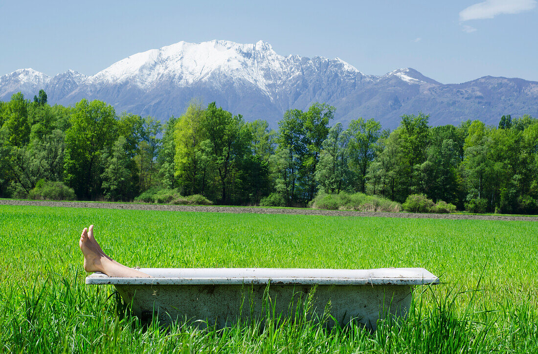 Feet Sticking Out Of A Bathtub In The Middle Of A Field With The Mountains Of The Swiss Alps In The Background,Locarno,Ticino,Switzerland