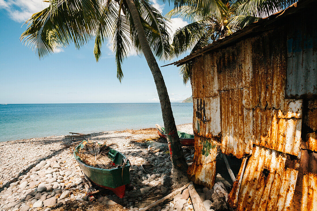 Blick auf den Ozean von der felsigen Strandpromenade mit einer Palme, einer verrosteten Fischerhütte und traditionellen Einbäumen, die entlang der Küste von Scotts Head in der Soufriere Bay auf der Insel Dominica gestrandet sind,Soufriere,Dominica,Karibik