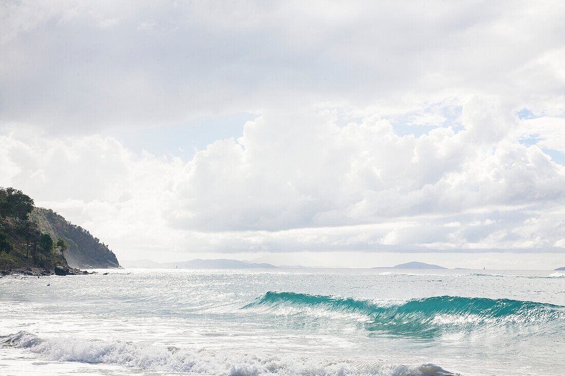 Türkisfarbene Welle und schäumende Brandung, die gegen einen blassgrauen, wolkenverhangenen Himmel am Strand von Cane Garden Bay, Tortola, Britische Jungferninseln, Karibik, ans Ufer rollt