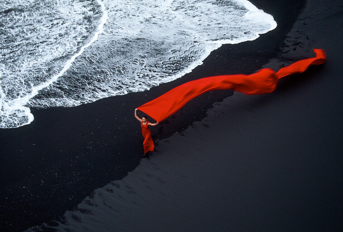 Woman in flowing red on a black sand beach at surf's edge in Iceland,Iceland