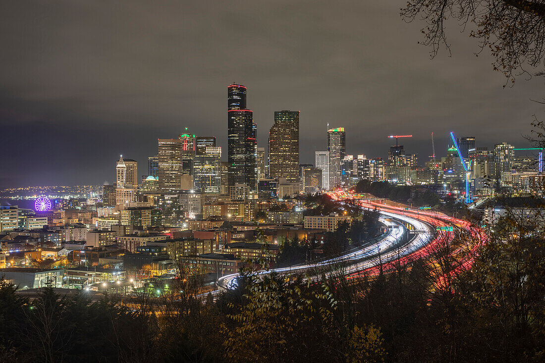 Nächtlicher Blick auf die Skyline von Seattle mit Verkehr auf der I-5 und dem Seattle Great Wheel ganz links, Seattle, Washington, Vereinigte Staaten von Amerika