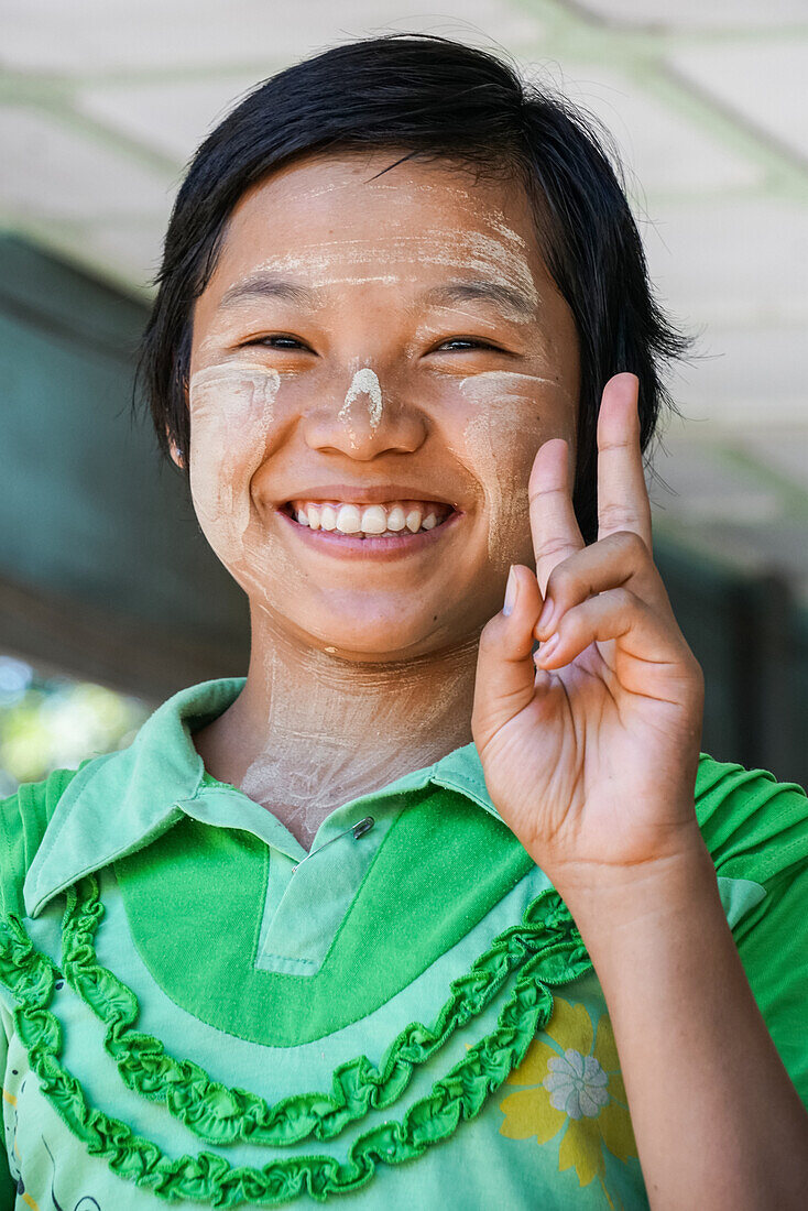 Burmesisches Mädchen mit Tananka-Paste, das die Kamera mit einem Friedenszeichen begrüßt, Myanmar