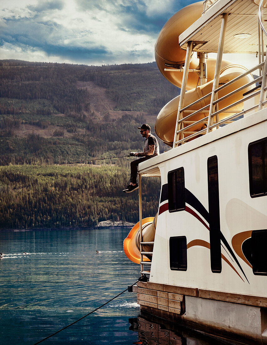 Man enjoying a houseboat,family vacation and fishing off the deck while parked on the shoreline of Shuswap Lake,Shuswap Lake,British Columbia,Canada