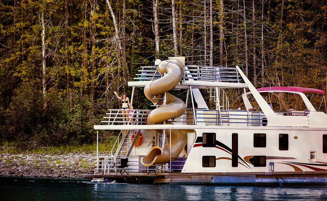 Eine Familie genießt ihren Urlaub auf einem Hausboot, das am Ufer des Shuswap Lake geparkt ist. Mutter und Tochter winken vom Deck am Heck des Bootes und ein Junge im Vorschulalter sitzt auf dem Deck im Hintergrund, Shuswap Lake, British Columbia, Kanada