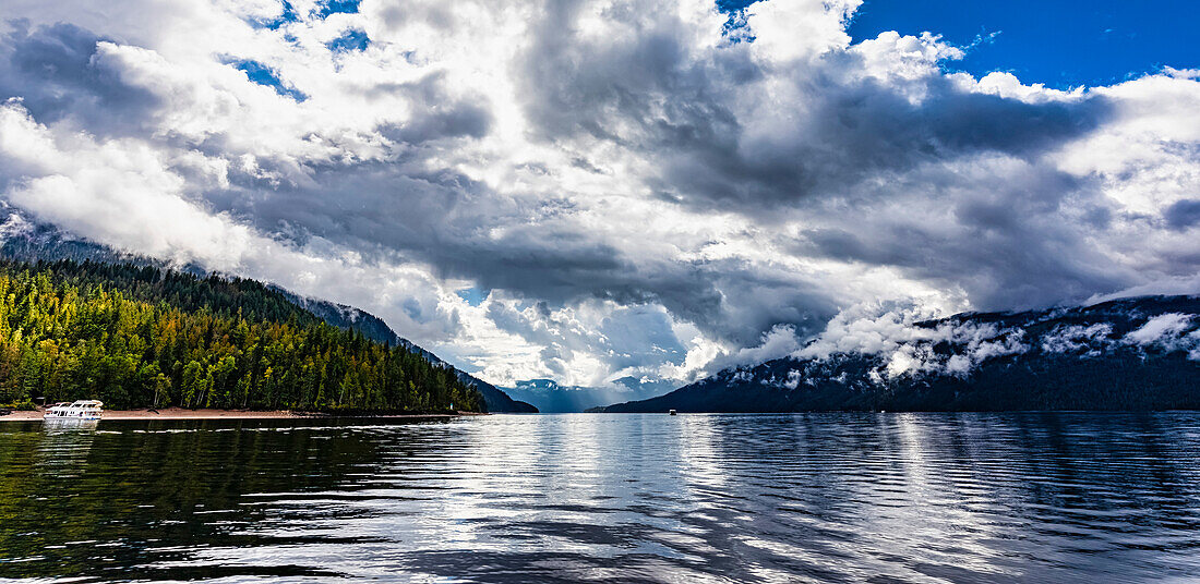 Landschaftlicher Blick auf den schönen Shuswap Lake mit dramatischen Wolken am Himmel, einem am Ufer vertäuten Hausboot und einem Boot auf dem Wasser während der Herbstsaison, Shuswap Lake, British Columbia, Kanada