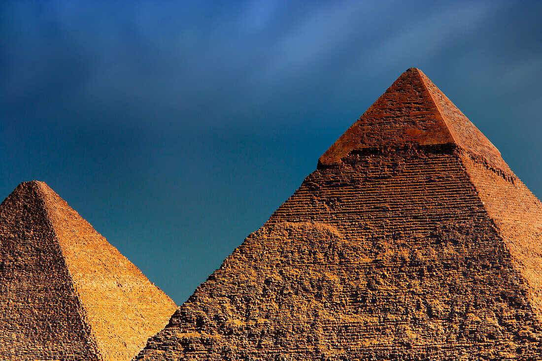 Zwei der Hauptpyramiden von Gizeh, von der Sonne mit einem goldenen Farbton beleuchtet, die Große Pyramide (Cheops) unter einem dunklen, bewölkten Himmel, Gizeh, Ägypten