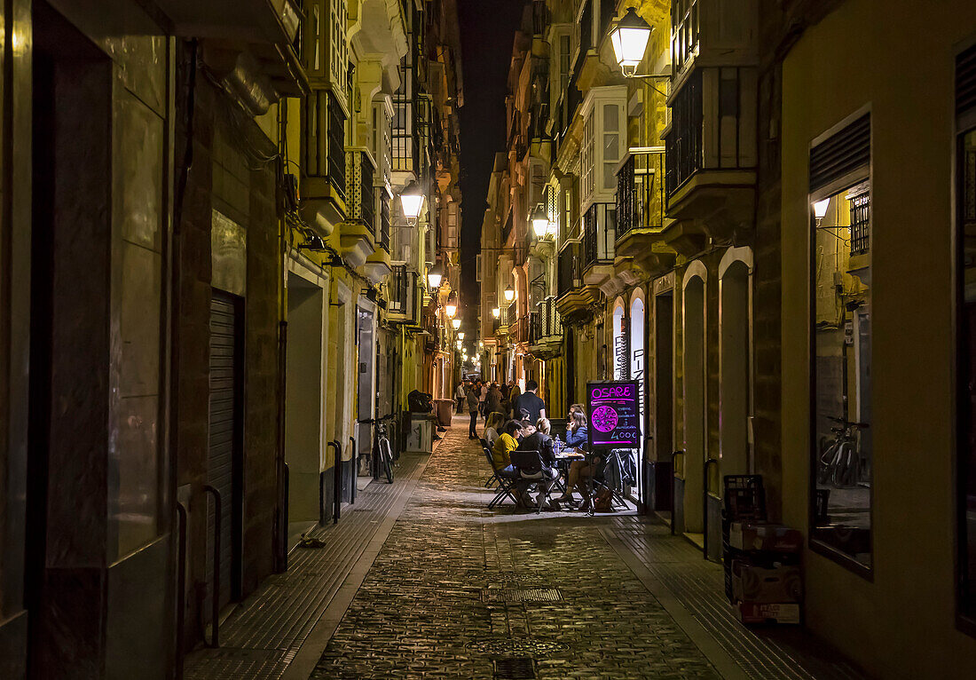 Gäste sitzen an Tischen in einem lichtdurchfluteten Innenhof in einer ruhigen, engen Straße bei Nacht, Cadiz, Andalusien, Spanien