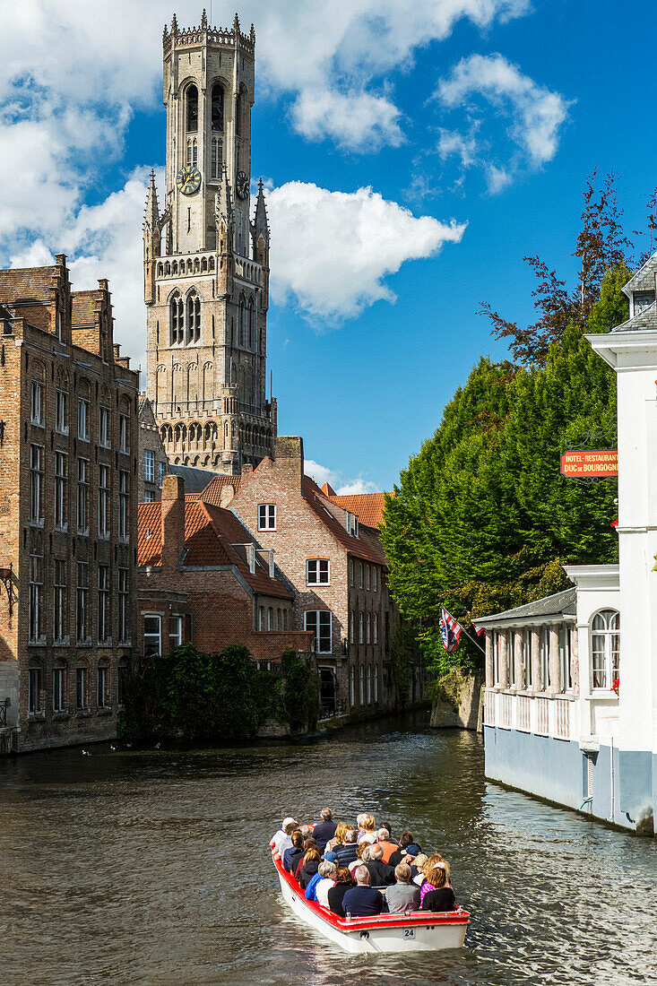 Kleines Ausflugsboot mit Menschen in einer Gracht mit großem gotischen Kirchturm im Hintergrund, Brügge, Belgien