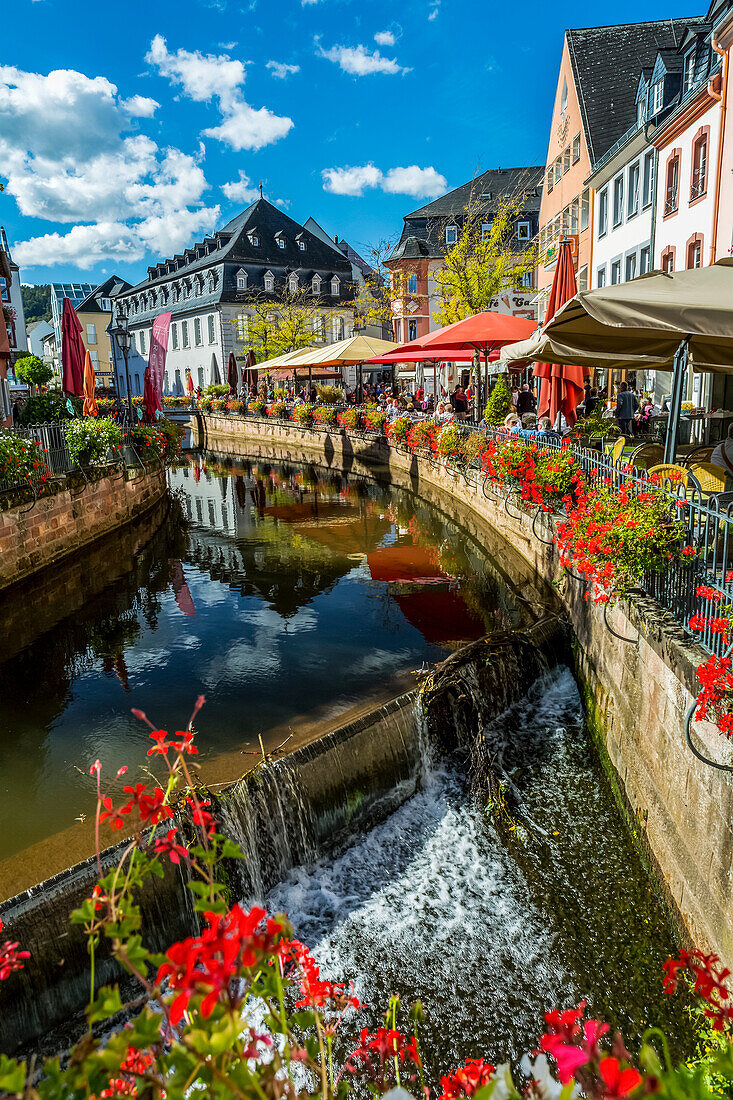 Kanal mit Wasserfällen in einem bunten Dorf mit Blumenkästen entlang des Geländers und blauem Himmel mit Wolken, Saarburg, Deutschland