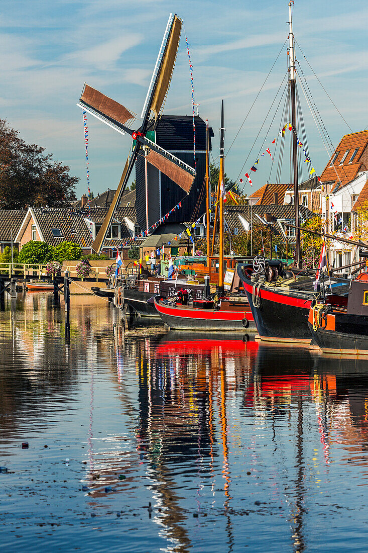Alte hölzerne Windmühle und alte Holzboote, die sich in einer Gracht spiegeln, eingerahmt von Gebäuden, blauem Himmel und Wolken, Leiden, Niederlande