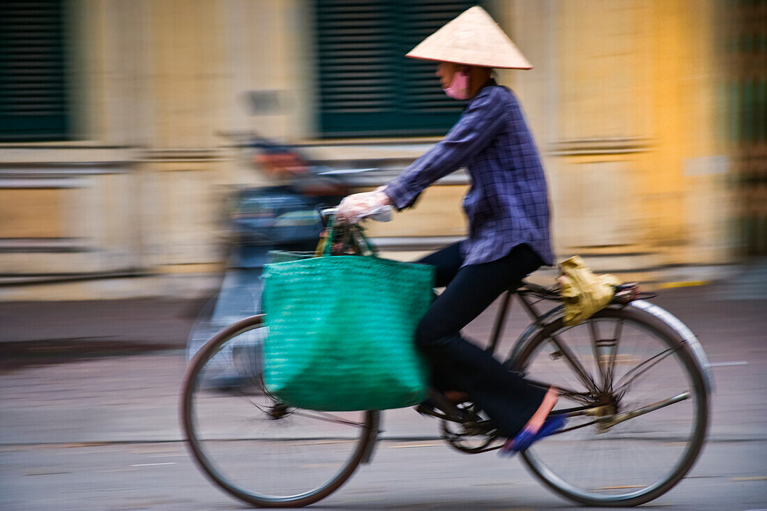 Bewegungsunschärfe einer Frau mit kegelförmigem Hut, die mit einer Einkaufstasche auf dem Fahrrad die Straße entlang fährt, Hanoi, Vietnam