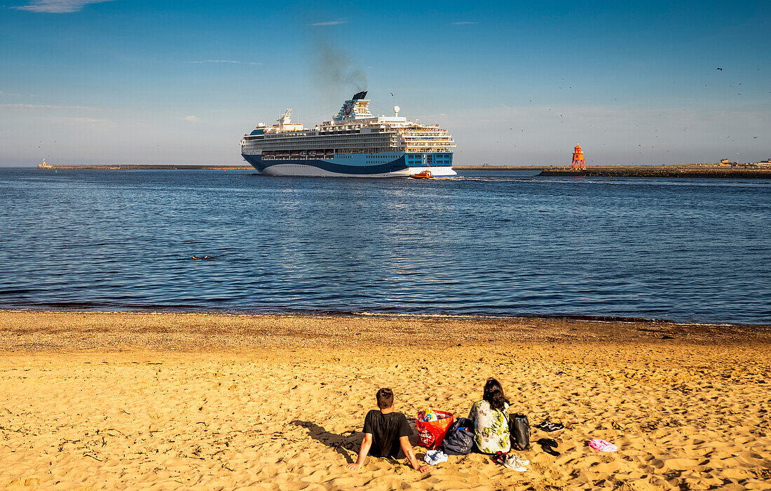 Ein Paar sitzt am Strand und beobachtet ein auslaufendes Kreuzfahrtschiff neben dem Herd Groyne Lighthouse, North Shields, Tyne and Wear, England