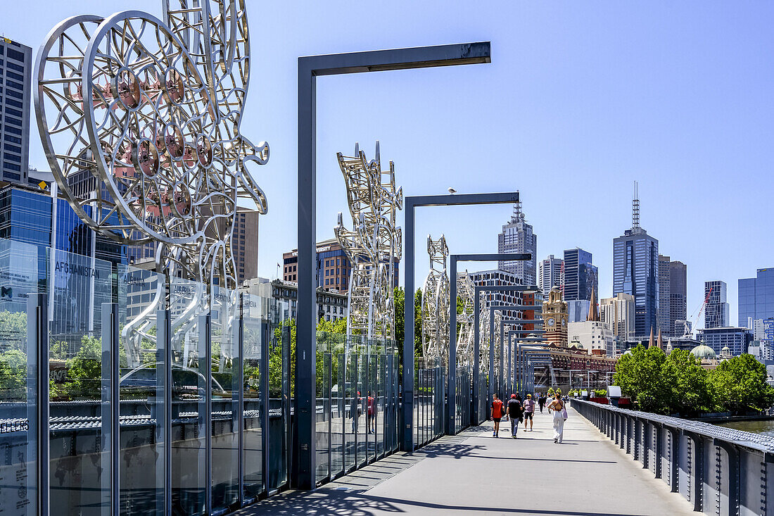 Skyline von Gebäuden in Melbourne von der Sandridge Bridge aus gesehen, die den Yarra River überquert, mit Skulpturen entlang der Brücke und Fußgängern, die in Richtung des Ufers gehen, Melbourne, Victoria, Australien