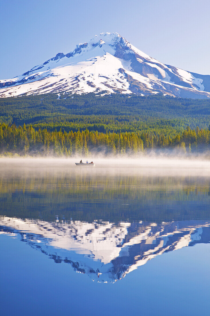 Mirror image of Mount Hood and forest reflected in Trillium Lake with mist rising off the water and people boating and fishing in the tranquil water,Oregon,United States of America