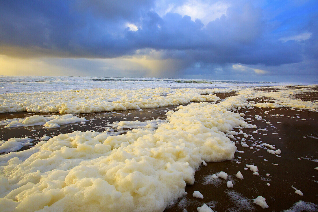 Meeresschaum am Strand mit anrollenden Wellen der Brandung unter einem stürmischen Himmel bei Sonnenuntergang, Oregon, Vereinigte Staaten von Amerika