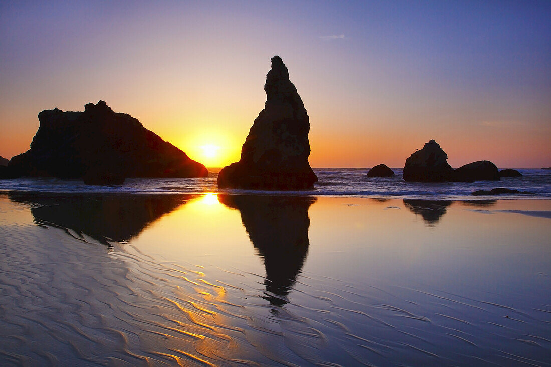 Zerklüftete Felsformationen entlang der Küste mit einer hellen Sonne, die bei Sonnenaufgang am Himmel leuchtet und sich auf dem nassen Sand in der Bandon State Natural Area an der Küste von Oregon spiegelt, Bandon, Oregon, Vereinigte Staaten von Amerika