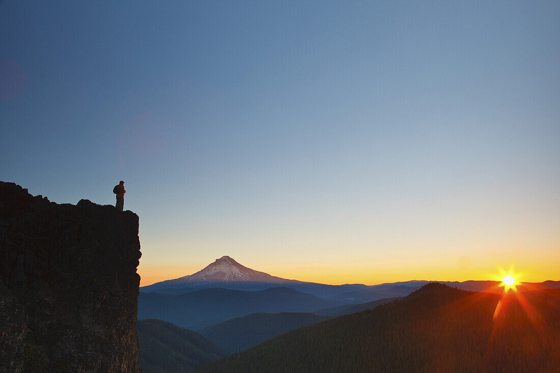 A man stands on a ridge looking out at Mount Hood with a sunburst over the horizon at sunrise,Oregon,United States of America