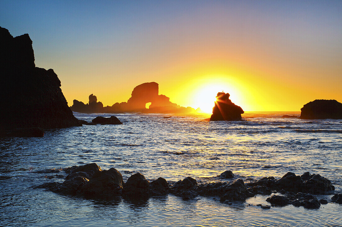 Helle untergehende Sonne, die unter dem Horizont über dem Wasser mit Felsformationen entlang der Küste von Oregon untergeht, Oregon, Vereinigte Staaten von Amerika