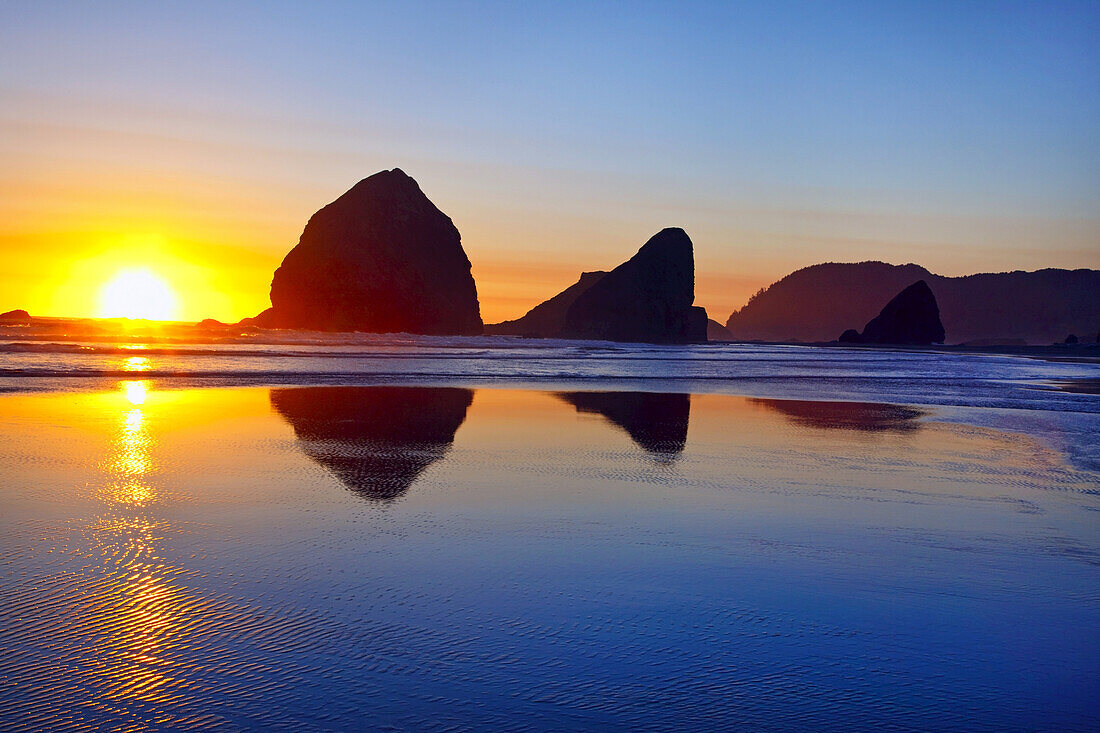 Felsformationen entlang des Strandes bei Sonnenuntergang im Cape Sebastian State Scenic Corridor. Die helle Sonne sinkt unter den Horizont und spiegelt sich im nassen Sand bei Ebbe, Oregon, Vereinigte Staaten von Amerika