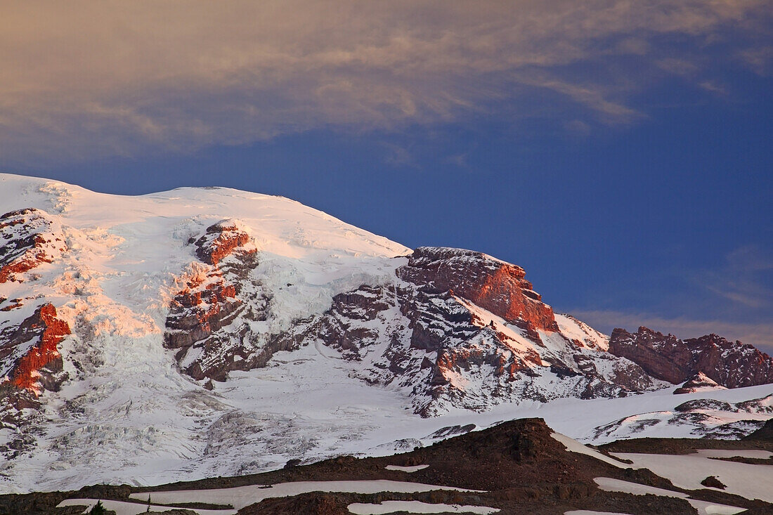 Schnee bedeckt die Felsen einer Gebirgskette bei Sonnenaufgang mit warmem Sonnenlicht, das den Gipfel beleuchtet, gesehen vom Paradise Park im Mount Rainier National Park, Washington, Vereinigte Staaten von Amerika